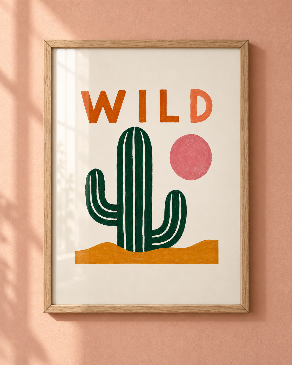 Framed desert-themed wall art hanging on a peach-pink wall, featuring the word “WILD” above a green cactus, a pink sun, and orange sand.