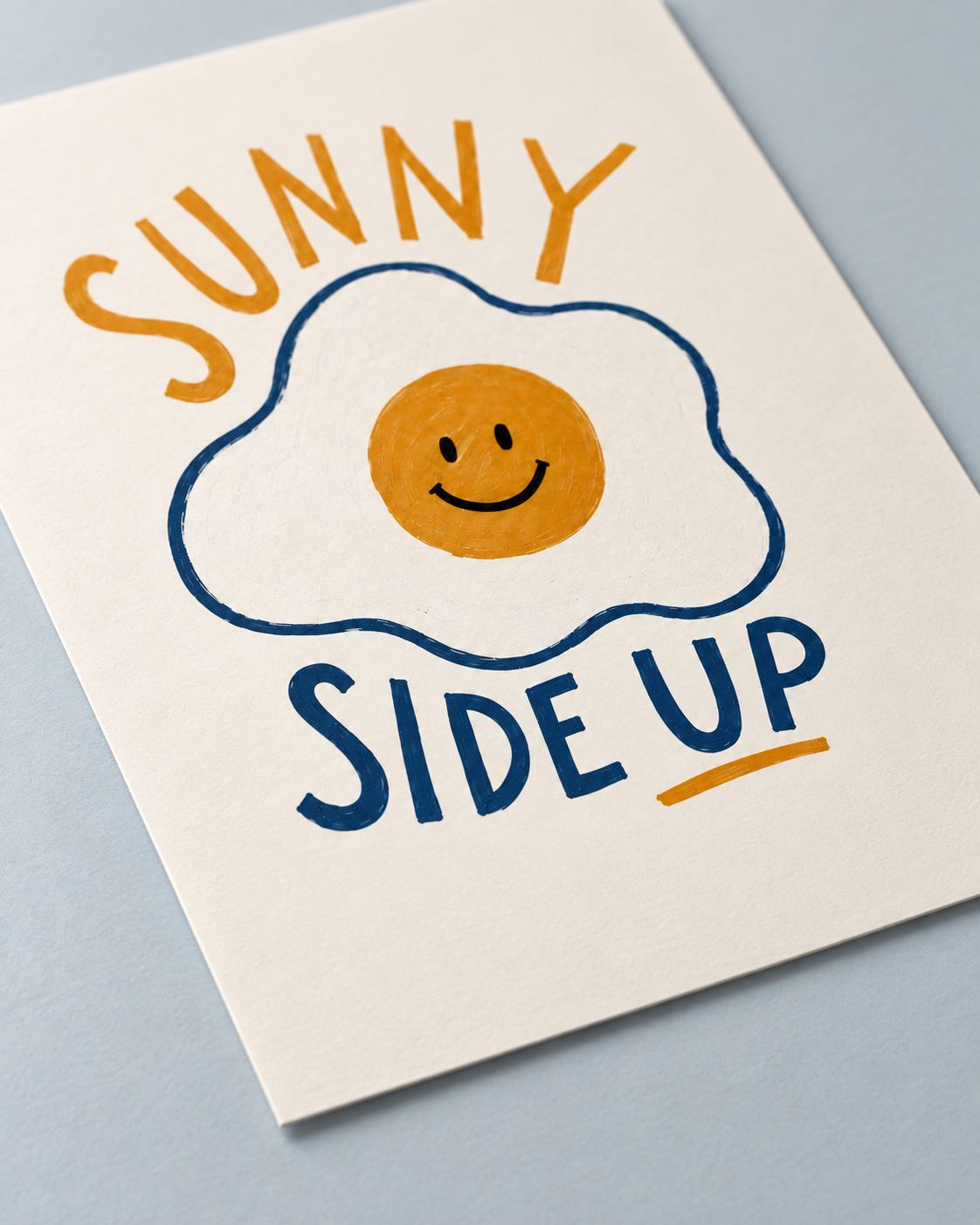 A square art print with a hand-drawn style design that reads 'SUNNY SIDE UP' in orange and navy blue lettering, surrounding a smiling fried egg illustration. The print is photographed at an angle on a light blue-gray surface.