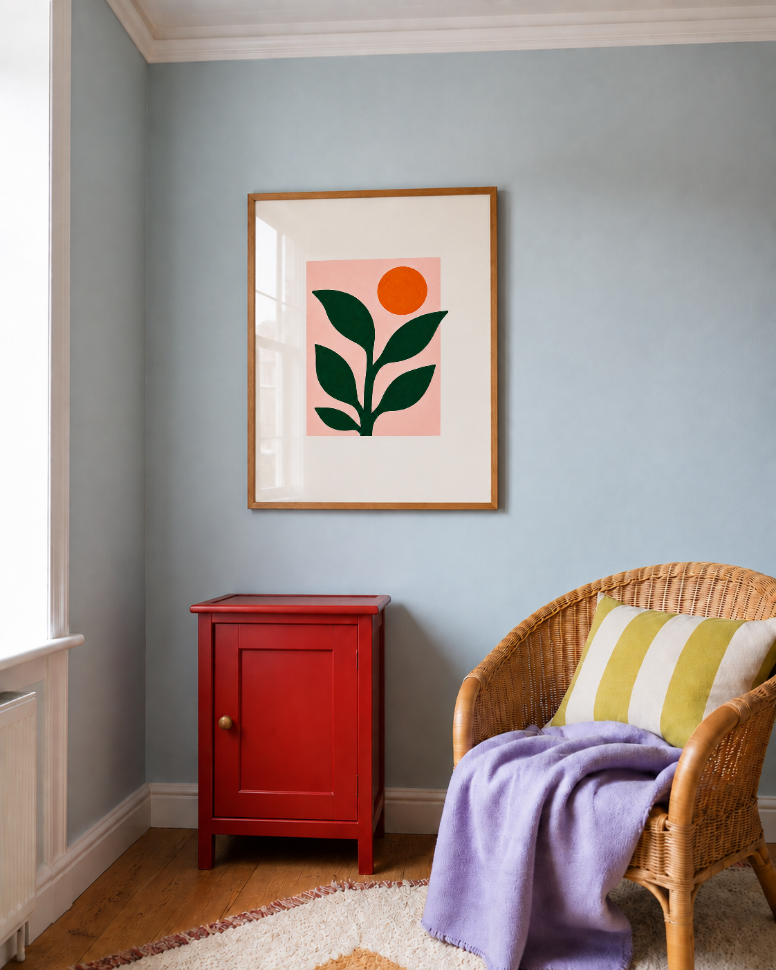 A framed minimalist art print featuring a green leaf and orange sun on a pink background, hung on a light blue-gray wall in a styled room with a red cabinet and a wicker armchair.