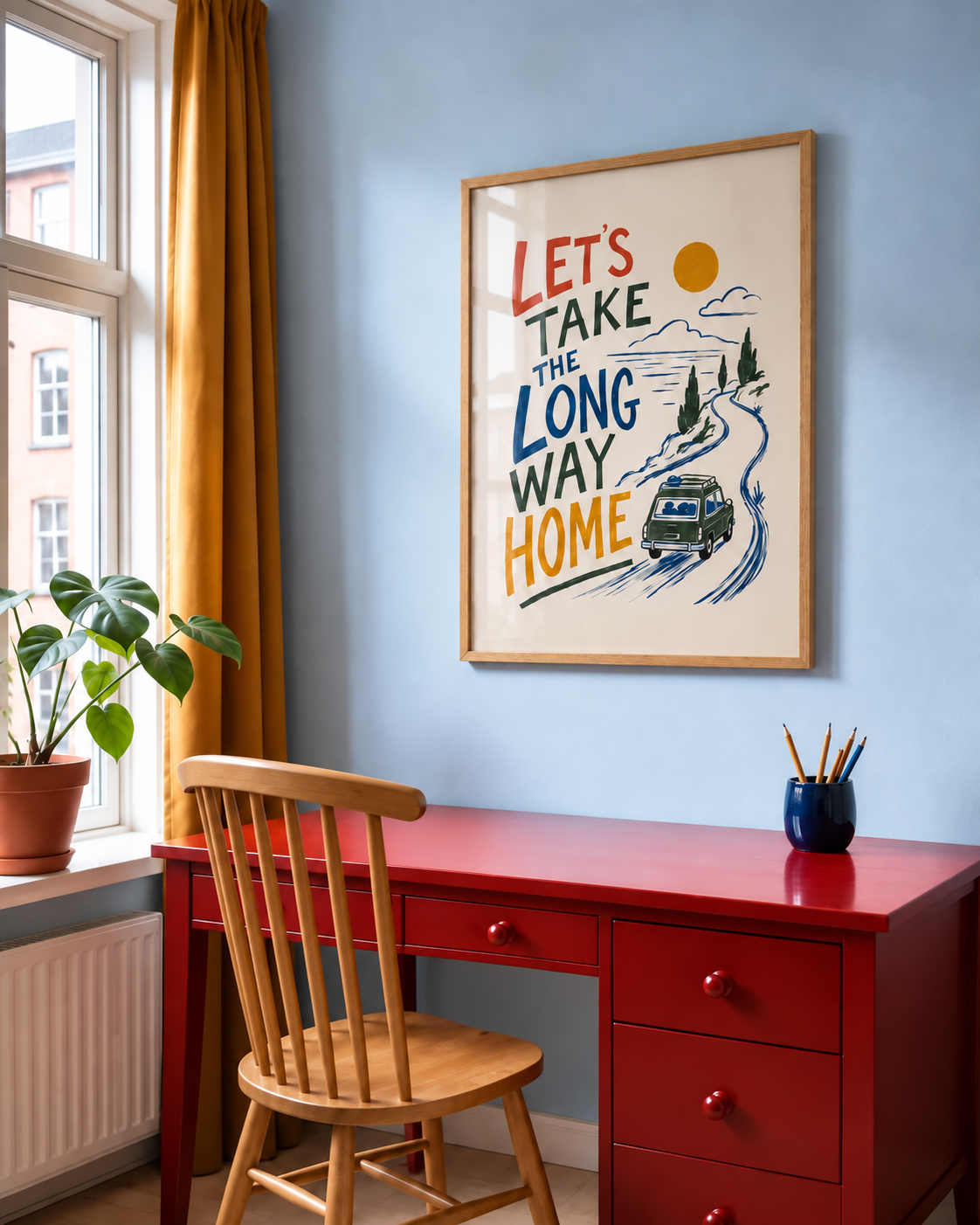 A framed art print with a vintage-style travel illustration and the text 'LET'S TAKE THE LONG WAY HOME' hangs on a light blue wall above a red wooden desk with a wooden chair in a styled home office.