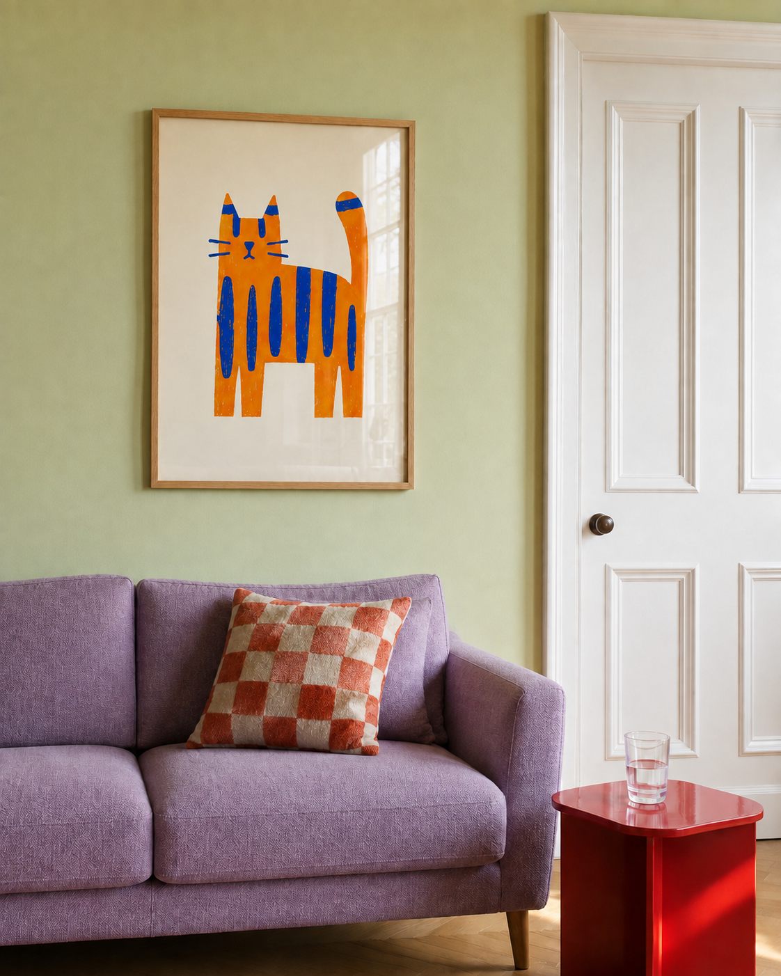 A framed art print of a stylized orange cat with blue stripes hanging on a sage green wall above a purple sofa with a checkered cushion.