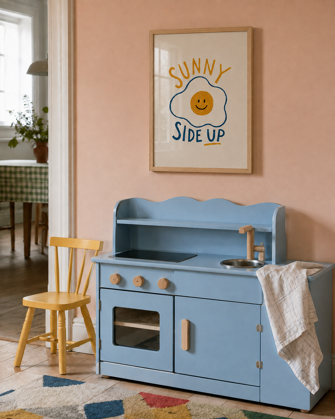 A light blue wooden toy play kitchen with a small sink, faucet, stove, and cabinet doors, styled in a nursery or children's room with a peach wall, framed 'Sunny Side Up' art, a yellow chair, and a patterned rug.