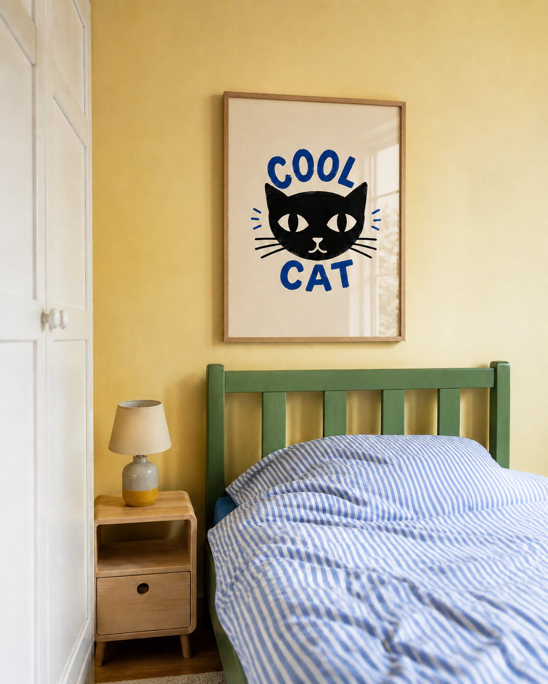 A framed wall art print featuring a black cat face with the words 'COOL CAT' in blue, hanging above a bed in a bedroom with a yellow wall, green headboard, and blue striped bedding.