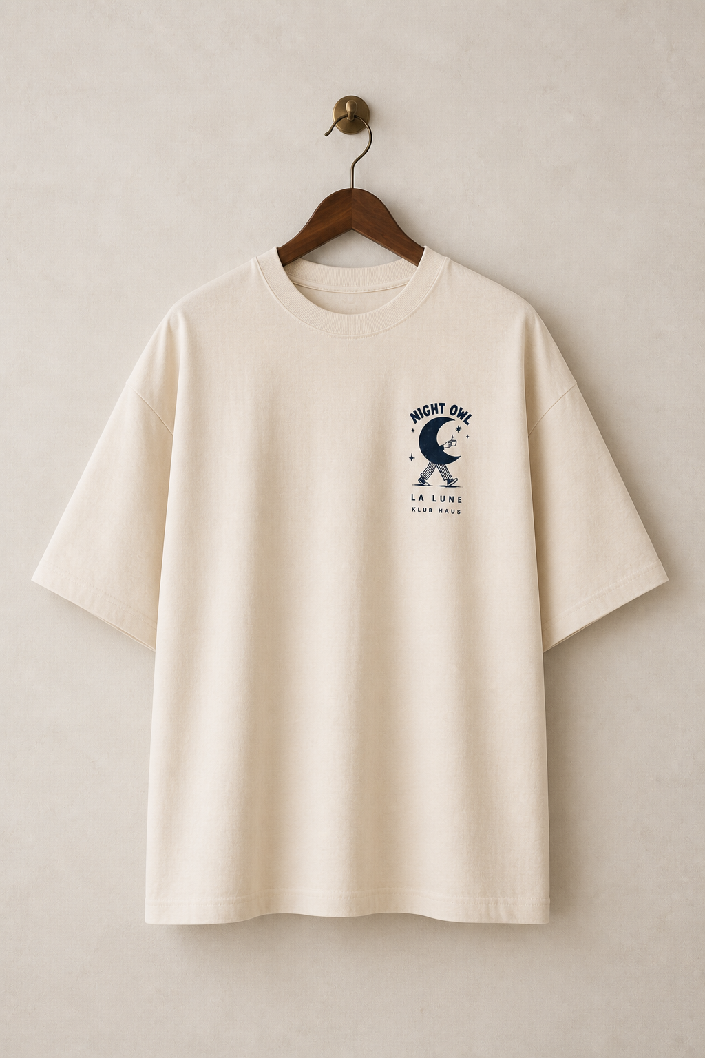 An off-white short-sleeve t-shirt hanging on a wooden hanger against a textured beige wall. The shirt features a dark navy blue graphic on the left chest depicting a crescent moon and a walking figure, with the text 'NIGHT OW' and 'LA LUNA' below it.