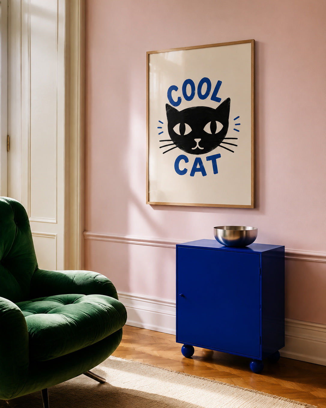 A framed wall art print featuring a black cat face with the words 'COOL CAT' in blue, hanging on a dusty pink wall in a styled room with a green armchair and a blue cabinet.