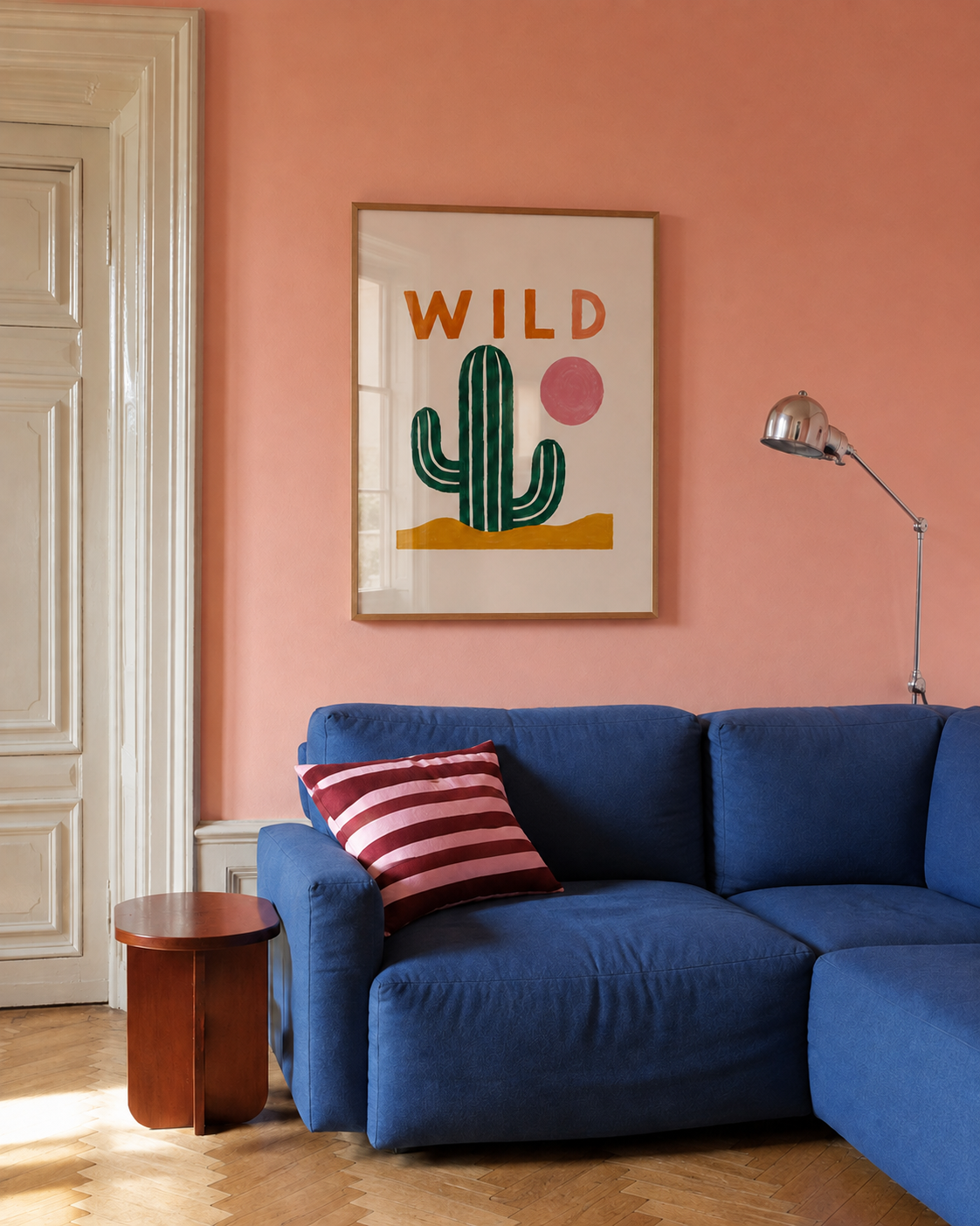 A framed art print featuring a green cactus and the word 'WILD' in orange, hanging on a terracotta-colored wall above a blue sectional sofa with a striped pillow.