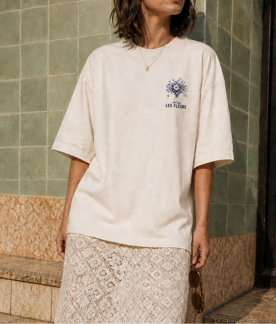 A woman wearing an oversized cream-coloured t-shirt with a blue graphic on the left chest and a matching cream lace midi skirt, standing on stone steps against a tiled wall.